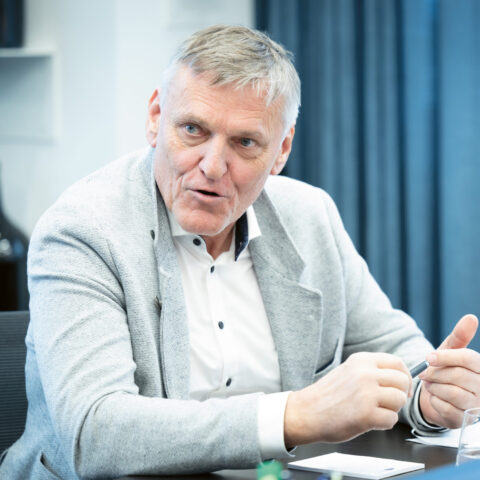 Ein älterer Mann in einem hellgrauen Blazer spricht, während er an einem Tisch sitzt und mit den Händen während einer Besprechung in einem modernen Büro gestikuliert.