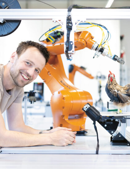 Ein lächelnder Mann lehnt an einem Tisch neben einem orangefarbenen Roboterarm und einem Modellhuhn in einer hellen Laborumgebung.