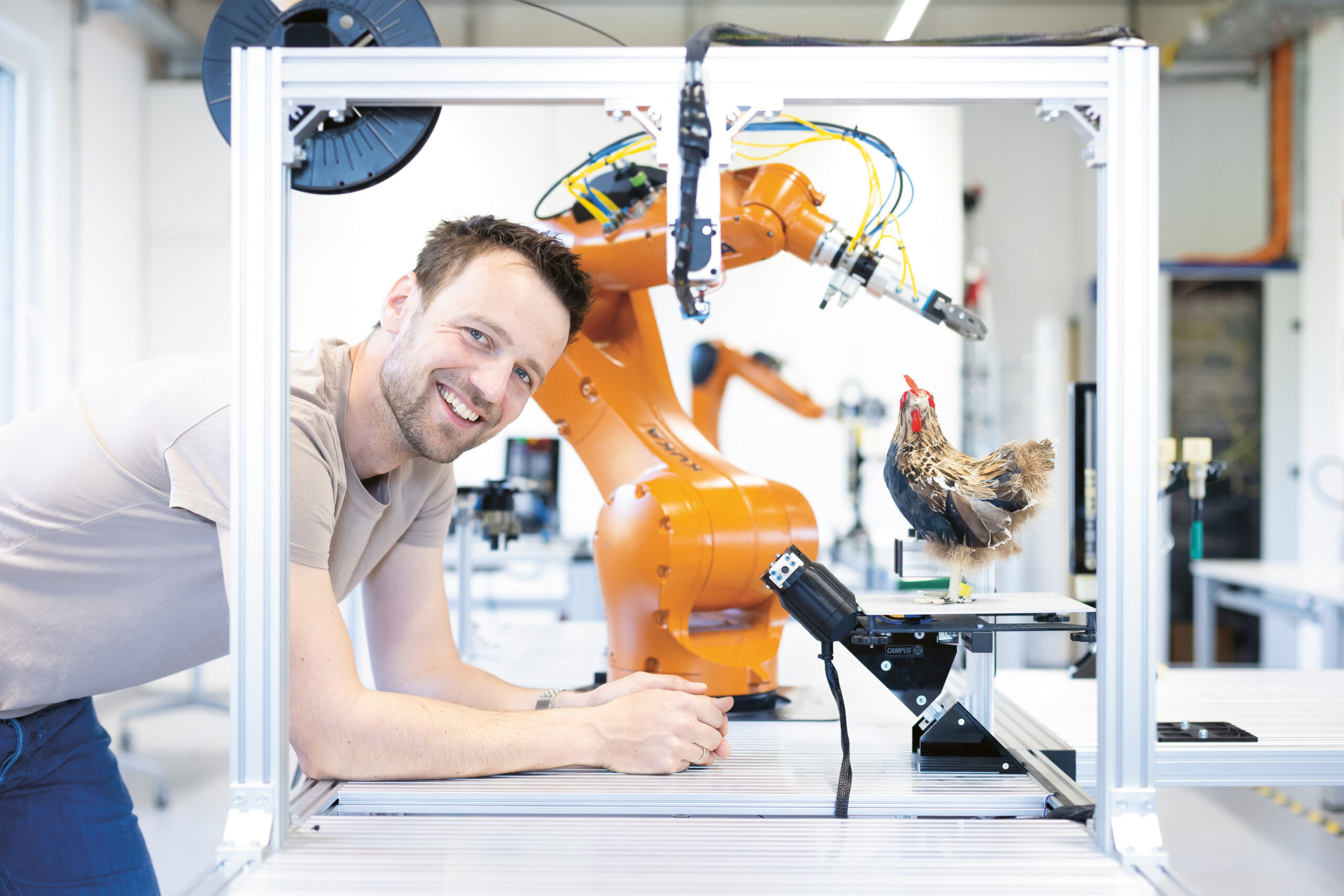 Ein lächelnder Mann lehnt an einem Tisch neben einem orangefarbenen Roboterarm und einem Modellhuhn in einer hellen Laborumgebung.