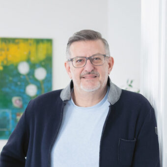 Zwei Männer mit Brillen und Jacken stehen in einem hellen, modernen Raum mit weißen Wänden und einem bunten Gemälde im Hintergrund.