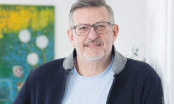 Zwei Männer mit Brillen und Jacken stehen in einem hellen, modernen Raum mit weißen Wänden und einem bunten Gemälde im Hintergrund.