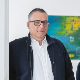 Zwei Männer in eleganter Freizeitkleidung stehen lächelnd in einem hellen Raum mit weißen Türen, Holzboden und bunter Kunst an der Wand.