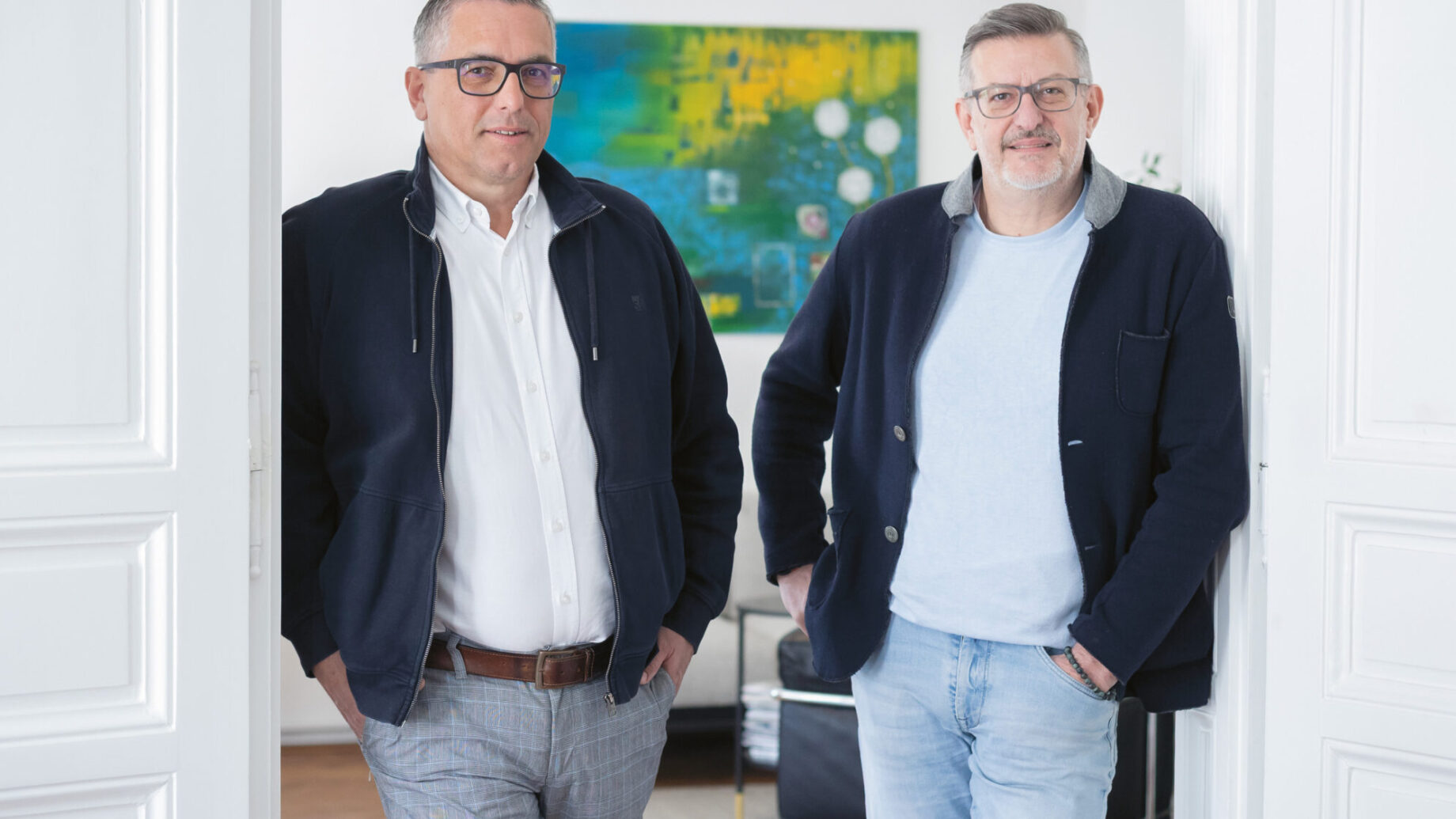 Zwei Männer mit Brillen und Jacken stehen lässig in einem hellen, modernen Raum mit weißen Wänden und einem bunten Gemälde im Hintergrund.