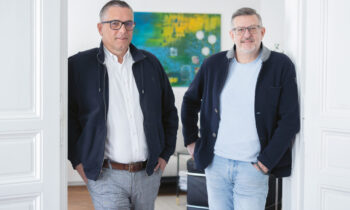 Zwei Männer mit Brillen und Jacken stehen lässig in einem hellen, modernen Raum mit weißen Wänden und einem bunten Gemälde im Hintergrund.