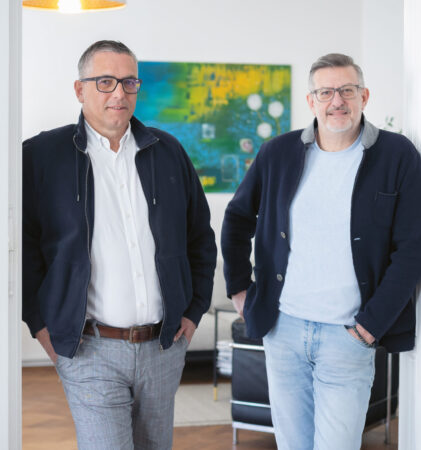 Zwei Männer mit Brillen und Jacken stehen lässig in einem hellen, modernen Raum mit weißen Wänden und einem bunten Gemälde im Hintergrund.