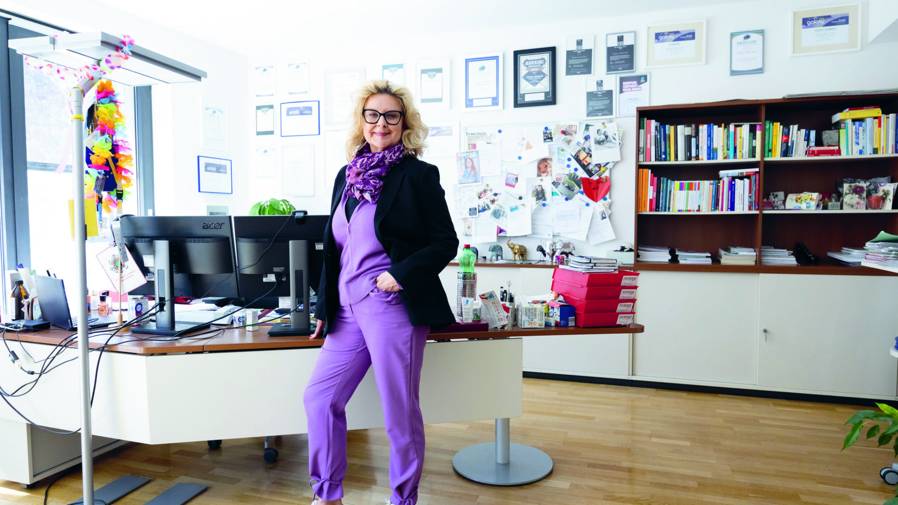 Eine Frau in lilafarbenem Outfit steht selbstbewusst in einem hellen, modernen Büro mit Bücherregalen, Urkunden und Papieren an der Wand hinter ihr.