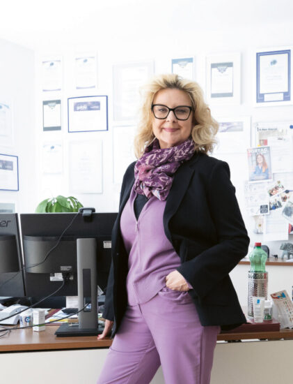 Eine Frau in einem lila Anzug steht selbstbewusst an ihrem Schreibtisch in einem hellen, modernen Büro mit Bücherregalen und Auszeichnungen an der Wand.