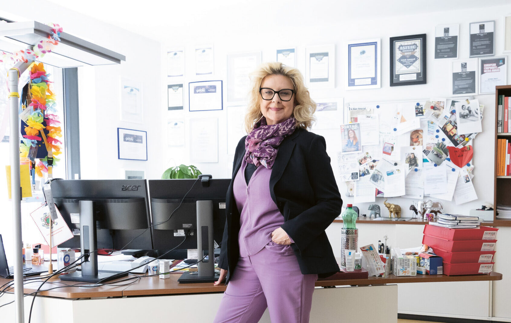 Eine Frau in einem lila Anzug steht selbstbewusst an ihrem Schreibtisch in einem hellen, modernen Büro mit Bücherregalen und Auszeichnungen an der Wand.