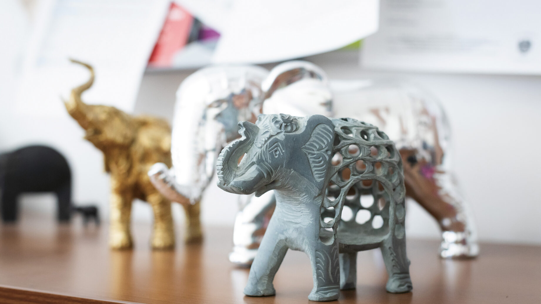 Drei dekorative Elefantenfiguren - eine graue und kunstvoll geschnitzte, eine goldene und eine silberne - stehen auf einem Holzregal mit Papieren im Hintergrund.