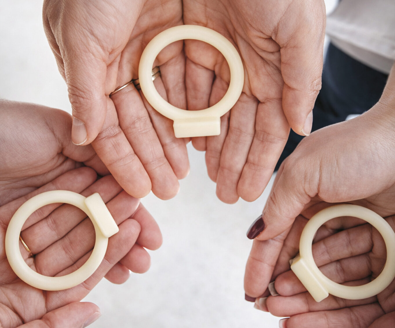 Vier Handpaare halten kreisförmige, blassgelbe Gegenstände, die jeweils einem Ring mit einer kleinen Lasche ähneln. Der Hintergrund ist unscharf.