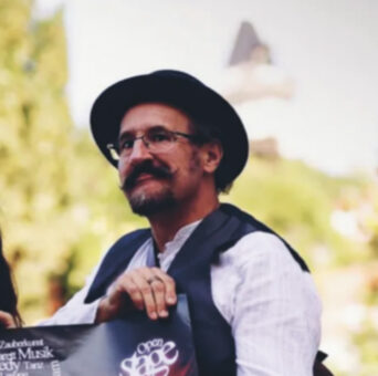Eine Frau und ein Mann in Vintage-Kleidung halten ein buntes Veranstaltungsplakat und Aufkleber auf einer Straße in der Stadt und lächeln in die Kamera.