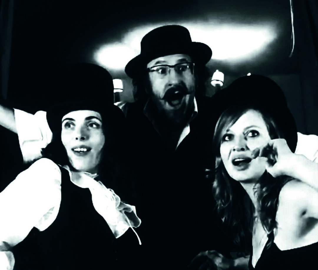 Drei Personen in Kostümen und Hüten im Vintage-Stil posieren dramatisch auf einem Schwarz-Weiß-Foto mit ausdrucksstarken Gesichtern und theatralischen Gesten.