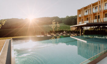 Modernes Hotel mit Liegestühlen am Außenpool, umgeben von grasbewachsenen Hügeln, bei Sonnenaufgang oder Sonnenuntergang mit Sonnenstrahlen.