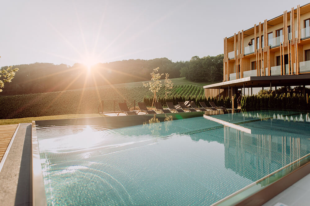Modernes Hotel mit Liegestühlen am Außenpool, umgeben von grasbewachsenen Hügeln, bei Sonnenaufgang oder Sonnenuntergang mit Sonnenstrahlen.