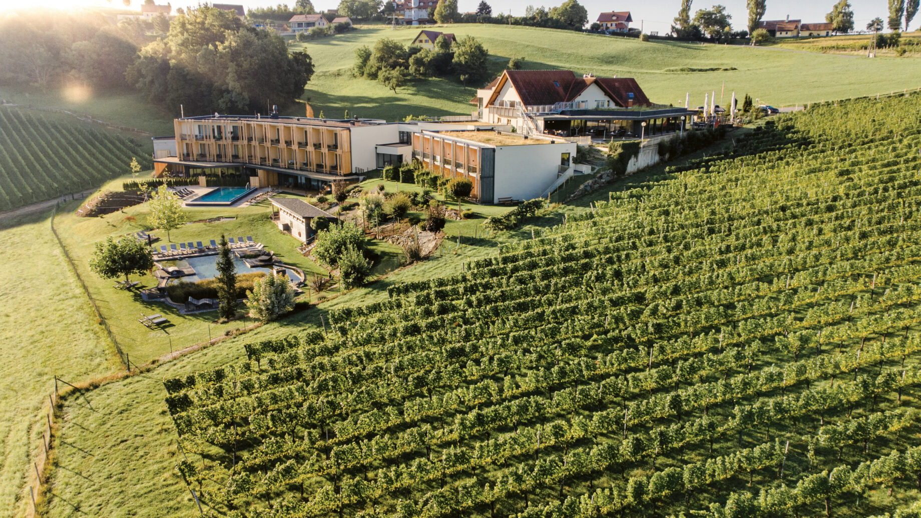 Ein modernes Hotel und ein Weinberg, umgeben von grünen Hügeln bei Sonnenaufgang, mit einem Pool, Gärten und entfernten Häusern im Hintergrund.