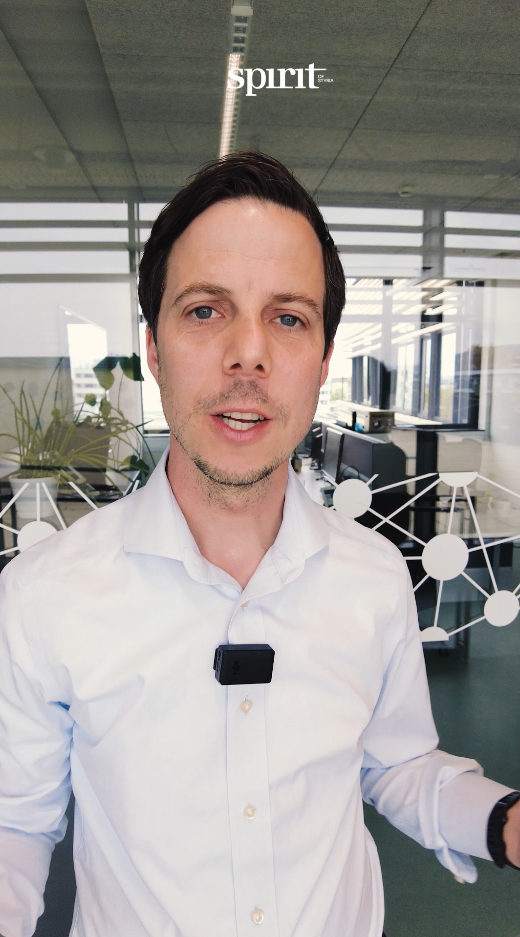 Ein Mann im weißen Hemd mit einem Mikrofon steht in einem modernen Büro