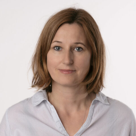 Frau mit schulterlangen hellbraunen Haaren, die ein helles Hemd zum Aufknöpfen trägt und in die Kamera schaut, vor einem schlichten weißen Hintergrund.