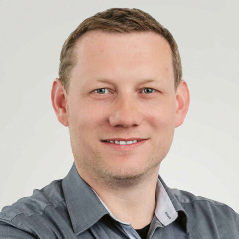 Lächelnder Mann mit kurzen hellbraunen Haaren, der ein graues Button-up-Hemd trägt und vor einem schlichten hellen Hintergrund steht.
