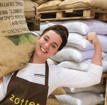 Zwei Personen lächeln in einer Lagerhalle, eine hält einen großen Sack auf der Schulter und trägt eine Schürze mit der Aufschrift "Zotter".