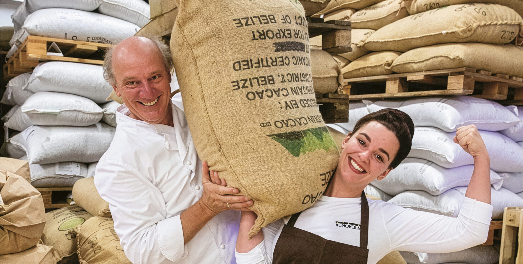 Eine lächelnde Frau in einer Zotter-Schürze beugt ihren Arm, während sie einen großen Sack aus Sackleinen auf ihrer Schulter hält; ein Mann steht neben ihr und hilft, den Sack zu stützen.