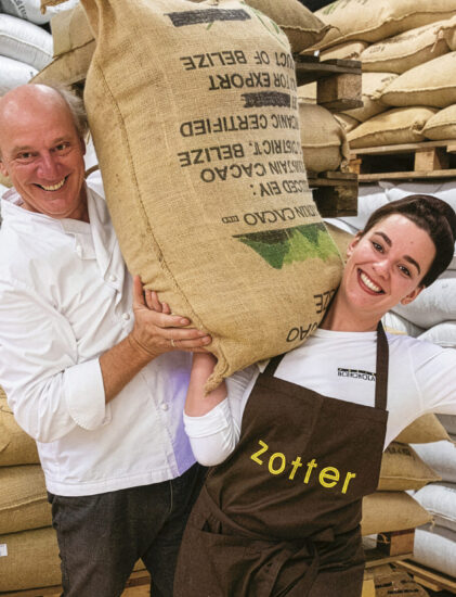 Eine lächelnde Frau in einer Zotter-Schürze beugt ihren Arm, während sie einen großen Sack aus Sackleinen auf ihrer Schulter hält; ein Mann steht neben ihr und hilft, den Sack zu stützen.