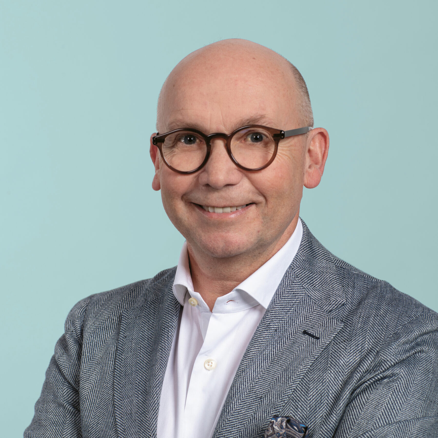 Glatzköpfiger Mann mit Brille, der einen grauen Blazer, ein weißes Hemd und eine Uhr trägt und mit verschränkten Armen vor einem hellblauen Hintergrund lächelt.