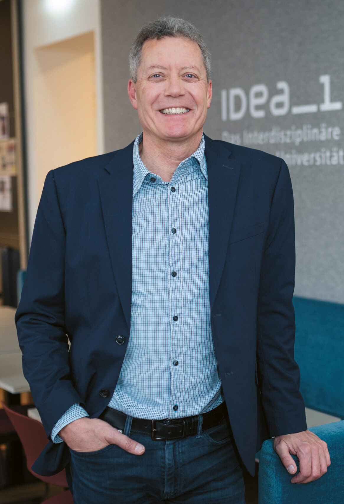 Ein lächelnder Mann im Blazer steht in einem modernen Büroraum mit "IDEA_Lab" und Universitätstext an der Wand hinter ihm.