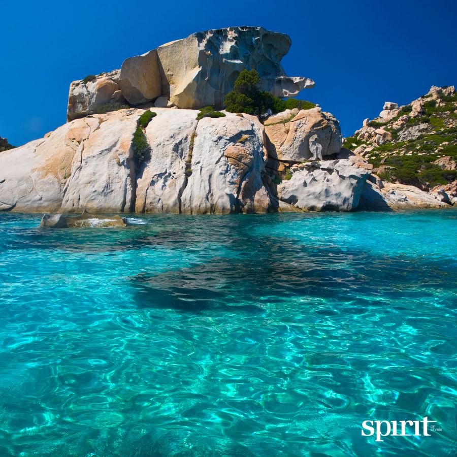 Eine felsige Klippe erhebt sich über türkisfarbenem Wasser unter blauem Himmel. In der rechten unteren Ecke steht in weißer Schrift "spirit" und GRAWE Bilanz 2025.