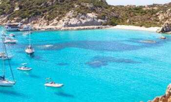 Türkisfarbene Bucht mit Segelbooten, Yachten, felsiger Küste und grünen Hügeln unter blauem Himmel. In der rechten unteren Ecke erscheint die GRAWE Bilanz 2025.