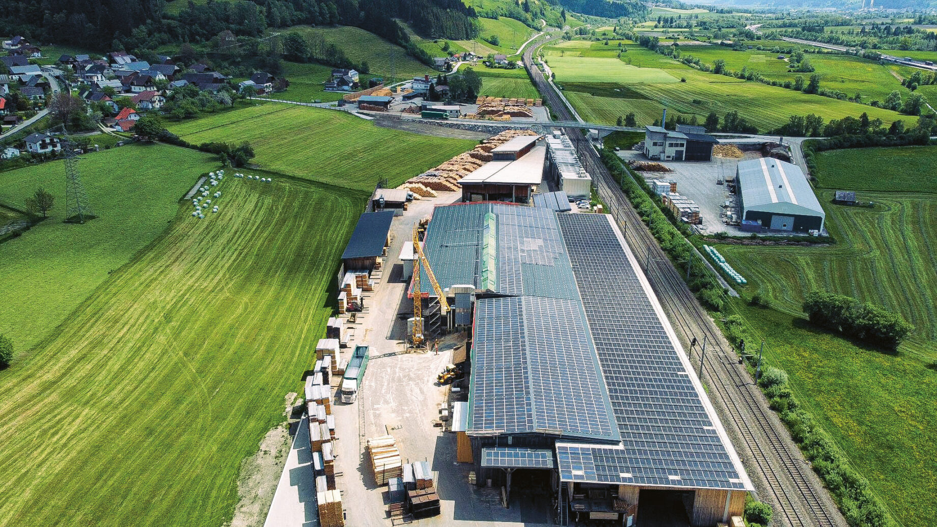 Luftaufnahme eines großen Industriegebäudes mit Sonnenkollektoren auf dem Dach, umgeben von grünen Feldern, Hügeln und einigen Gebäuden in der Nähe.