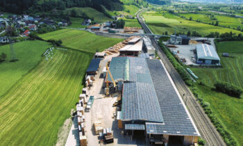 Luftaufnahme eines großen Industriegebäudes mit Sonnenkollektoren auf dem Dach, umgeben von grünen Feldern, Hügeln und einigen Gebäuden in der Nähe.