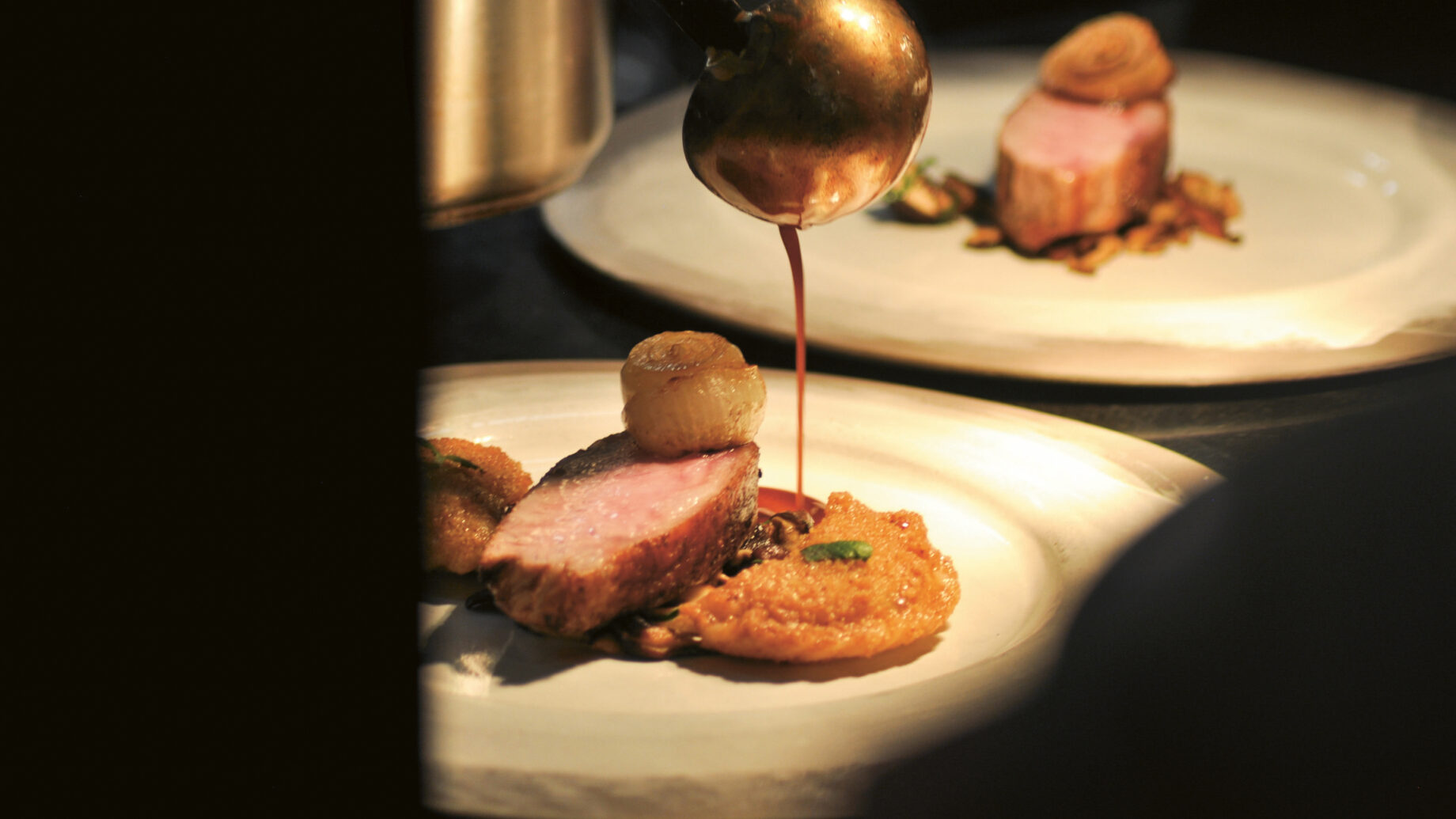 Ein Koch gießt Sauce über ein Gourmet-Gericht mit geschnittenem Fleisch, Gemüse und einem goldenen Püree auf einem weißen Teller.