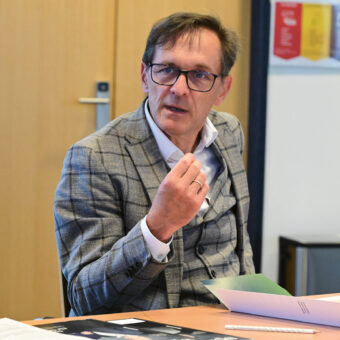 Ein Mann in einer karierten Anzugsjacke sitzt an einem Tisch mit Papieren und gestikuliert, während er in einem Büro oder Besprechungsraum spricht.