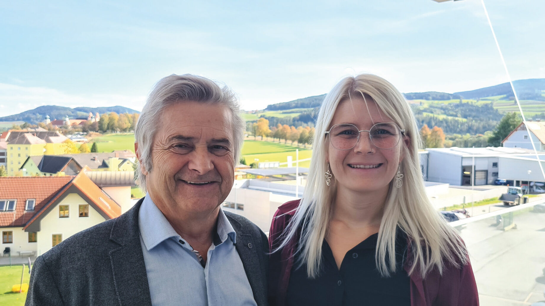 Ein älterer Mann und eine jüngere Frau stehen lächelnd im Freien, vor einer malerischen Kulisse aus Hügeln, Bäumen und Gebäuden unter blauem Himmel.