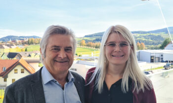 Ein älterer Mann und eine jüngere Frau stehen lächelnd im Freien, vor einer malerischen Kulisse aus Hügeln, Bäumen und Gebäuden unter blauem Himmel.