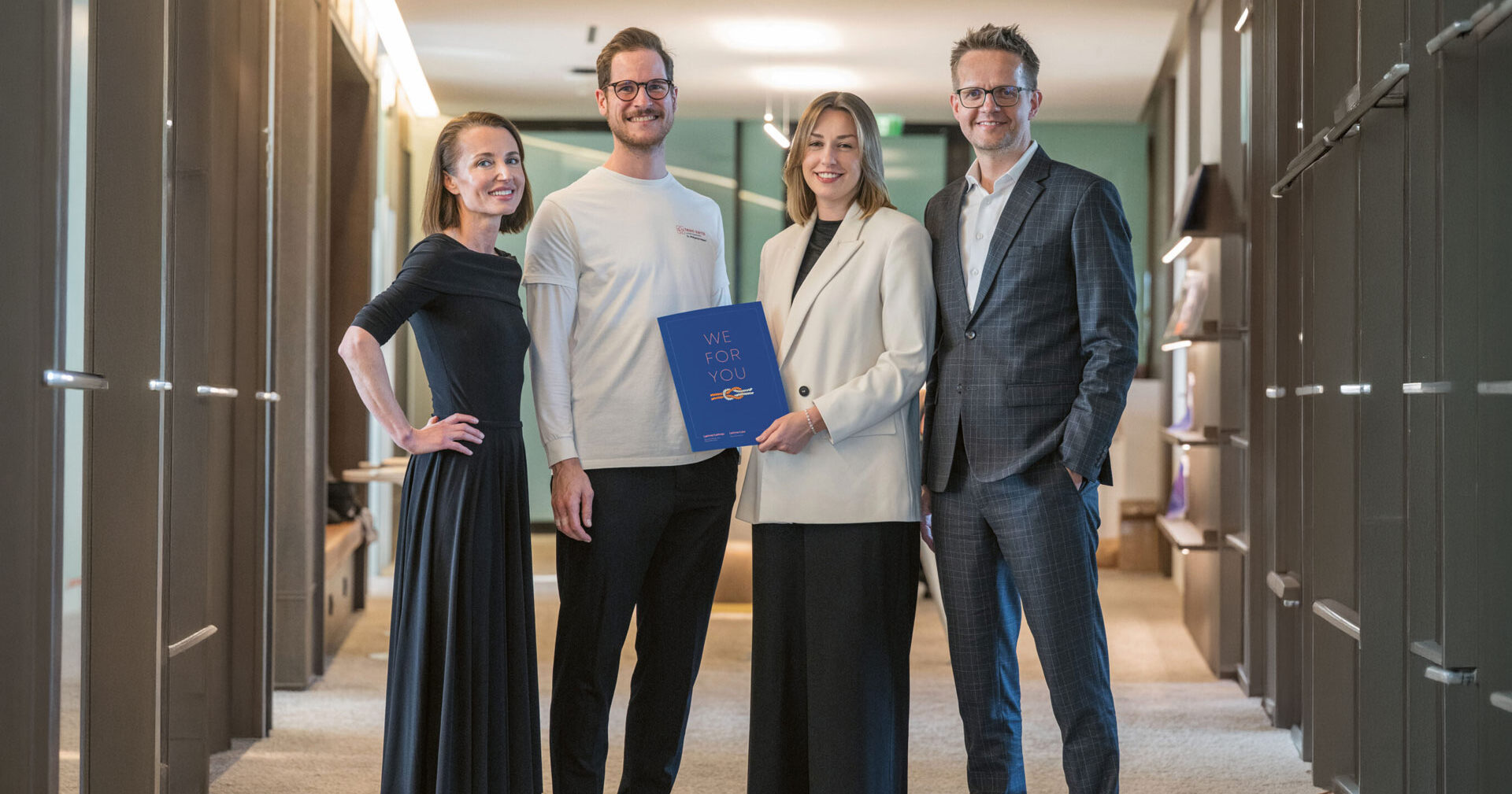 Vier professionell gekleidete Menschen stehen lächelnd in einem Büroflur; eine Frau hält einen blauen Ordner mit der Aufschrift "WIR FÜR SIE, SIE FÜR".