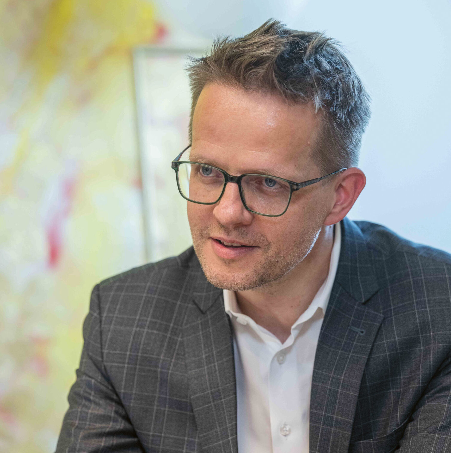 Ein Mann mit Brille und karierter Anzugsjacke sitzt an einem Schreibtisch, hält einen Stift in der Hand und spricht, während er ein Notizbuch vor sich liegen hat.