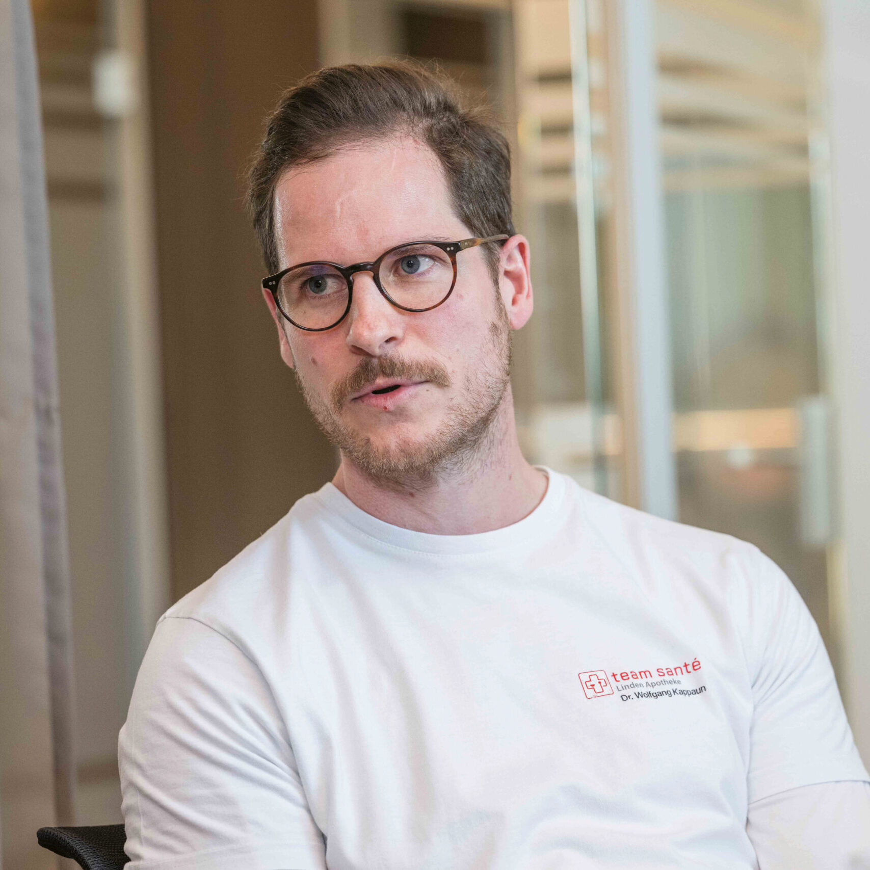 Ein Mann mit Brille und Bart, der ein weißes T-Shirt trägt, sitzt in einem Haus und schaut leicht zur Seite, als ob er sich unterhalten würde.