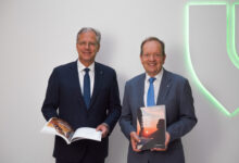 Zwei Männer in Anzügen stehen lächelnd mit aufgeschlagenen Büchern vor einem leuchtend grünen Logo - Prominenter Keynote Speaker im Grazer Membersclub J22: Peter Filzmaier.
