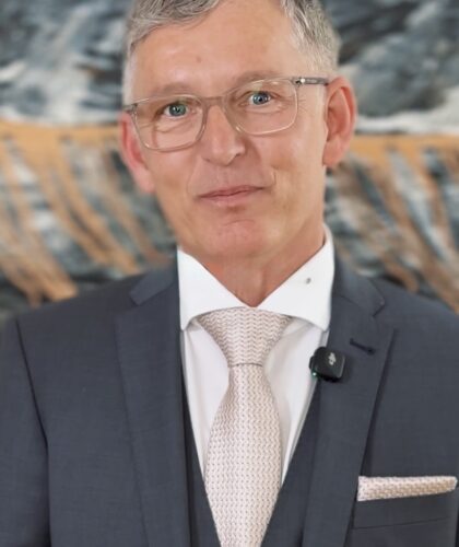 Ein Mann im grauen Anzug mit leichter Krawatte steht vor einem abstrakten Hintergrund.