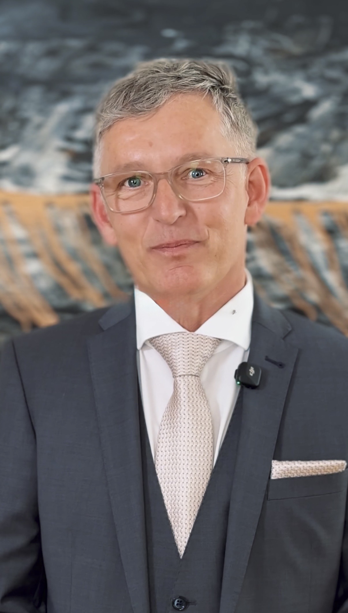 Ein Mann im grauen Anzug mit leichter Krawatte steht vor einem abstrakten Hintergrund.