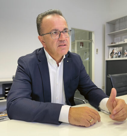 Ein Mann in einem marineblauen Anzug und mit Brille sitzt an einem weißen Schreibtisch und spricht mit der Hand, während er in einer modernen Büroumgebung spricht.