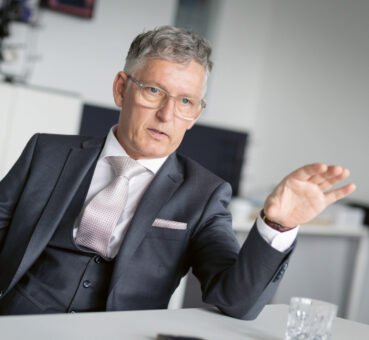 Ein Mann in Anzug und Krawatte sitzt an einem Schreibtisch und gestikuliert mit seiner rechten Hand während eines Gesprächs in einer modernen Büroumgebung.