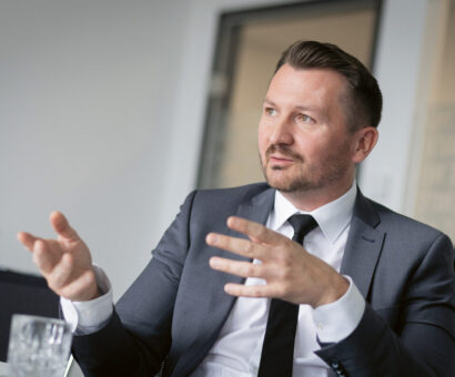 Ein Mann in Anzug und Krawatte gestikuliert mit seinen Händen, während er an einem Tisch in einem Büro sitzt.