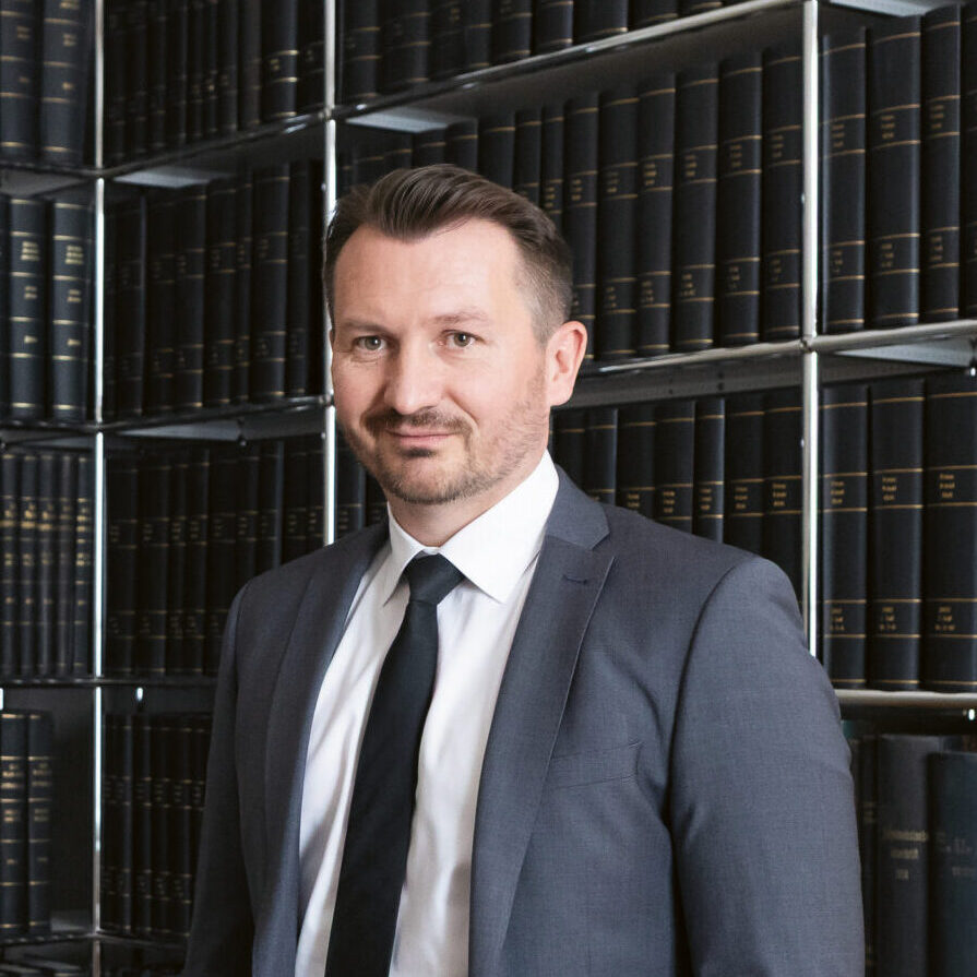 Zwei Männer in Anzügen stehen in einer Bibliothek mit Regalen voller schwarzer Bücher, posieren und lächeln in die Kamera.