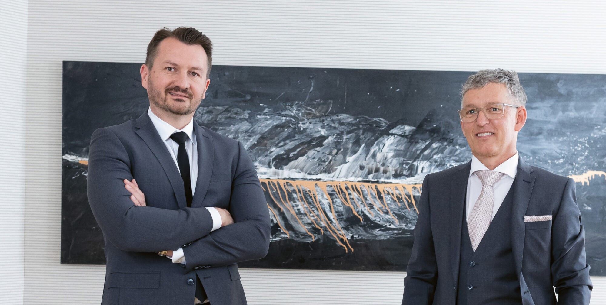 Zwei Männer in Anzügen stehen selbstbewusst in einem modernen Büro mit einem Konferenztisch, Stühlen und abstrakter Kunst an der Wand hinter ihnen.