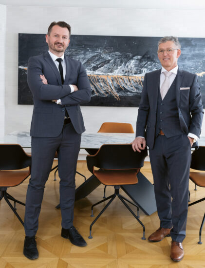 Zwei Männer in Anzügen stehen selbstbewusst in einem modernen Büro mit einem Konferenztisch, Stühlen und abstrakter Kunst an der Wand hinter ihnen.