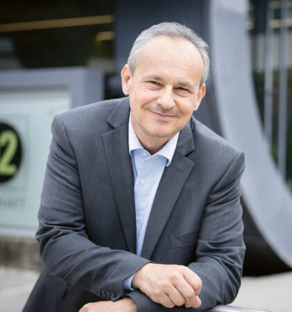 Ein Mann mittleren Alters im grauen Anzug lehnt lächelnd an einem Geländer vor einem modernen Gebäude mit Glasscheiben und einem Schild mit der Aufschrift "02.
