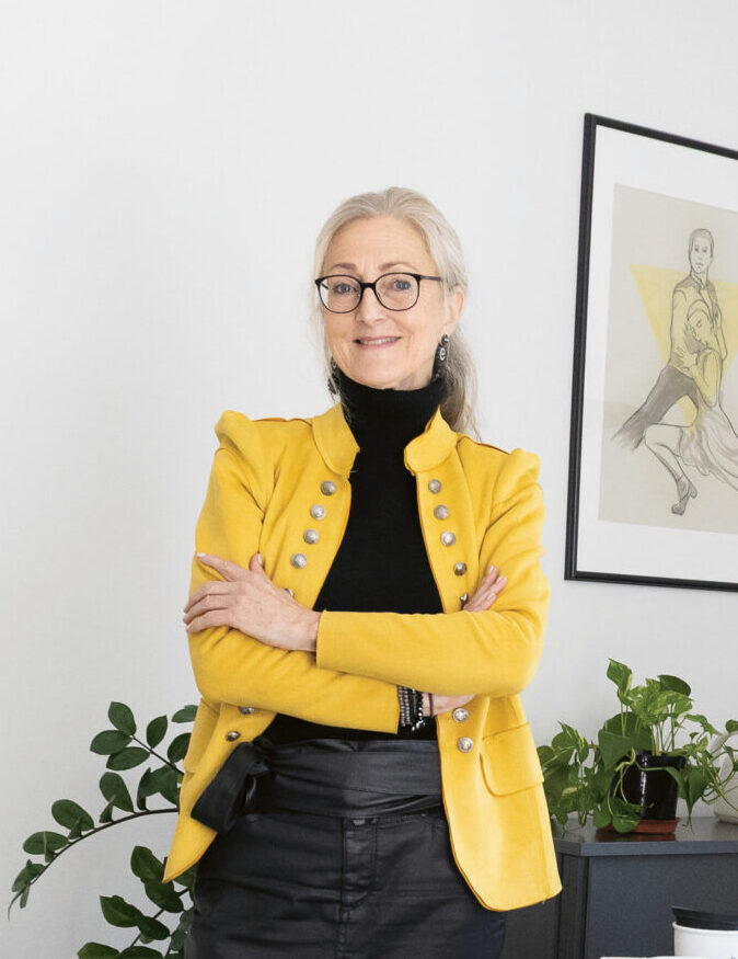 Eine Frau in einem gelben Blazer steht selbstbewusst in einem modernen Büro neben einem Schreibtisch mit Büchern, einem Computer und Büromaterial.
