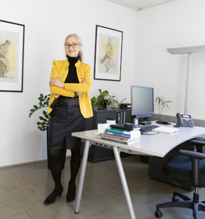 Eine Frau in einem gelben Blazer steht selbstbewusst an ihrem Schreibtisch, an dem Bücher, ein Computer und Pflanzen stehen; an den weißen Wänden hängt Kunst.